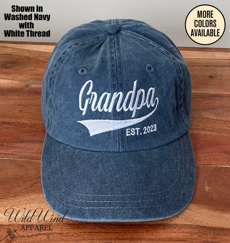 May include: A blue washed cotton baseball cap with white embroidery that reads "Grandpa EST. 2023".