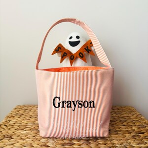 May include: A Halloween-themed tote bag with orange and white vertical stripes. The bag has a handle and the name "Grayson" printed on the front. Inside the bag is a white ghost plush toy wearing a "Spook" banner.