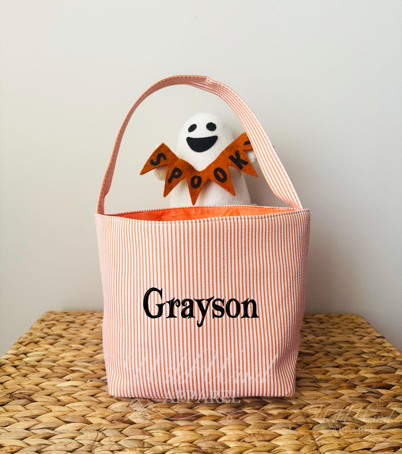 May include: A Halloween-themed tote bag with orange and white vertical stripes. The bag has a handle and the name "Grayson" printed on the front. Inside the bag is a white ghost plush toy wearing a "Spook" banner.