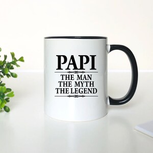 May include: White ceramic coffee mug with a black rim and handle. The mug has black text that reads "Papi The Man The Myth The Legend".