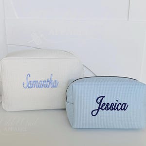 May include: Two waffle-textured cosmetic bags. One is white with "Samantha" embroidered in blue. The other is light blue with "Jessica" embroidered in navy. Both have a zipper closure and a small handle.