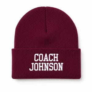 May include: A deep burgundy knit beanie featuring a folded cuff. The front of the cuff displays the text COACH JOHNSON embroidered in bold, white, block-style capital letters centered on the fabric.