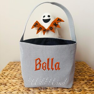 May include: A Halloween-themed tote bag with black and white stripes, featuring the name "Bella" embroidered in orange. A white ghost with an orange "Spooky" banner is inside the bag. The bag has a striped handle.