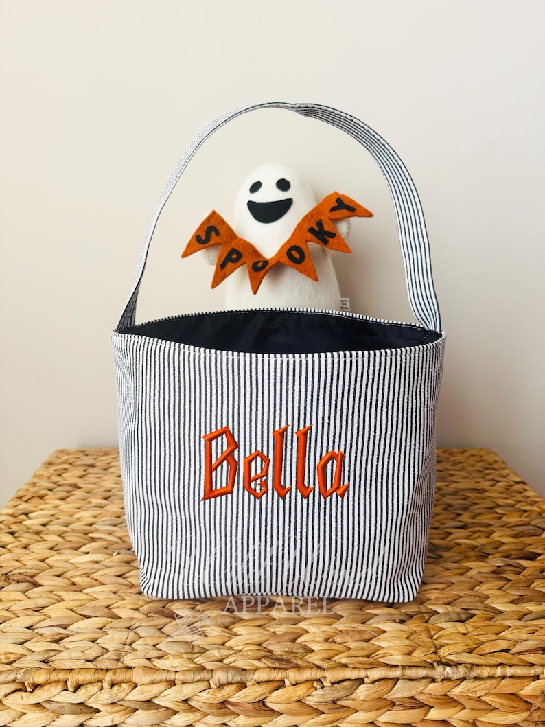 May include: A Halloween-themed tote bag with black and white stripes, featuring the name "Bella" embroidered in orange. A white ghost with an orange "Spooky" banner is inside the bag. The bag has a striped handle.