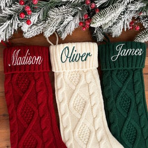 May include: Three knit Christmas stockings in red, cream, and dark green, each featuring a cable knit pattern and a folded cuff. The red stocking is embroidered with the name Madison, the cream with Oliver, and the green with James.