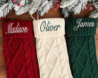 May include: Three knit Christmas stockings in red, cream, and dark green, each featuring a cable knit pattern and a folded cuff. The red stocking is embroidered with the name Madison, the cream with Oliver, and the green with James.