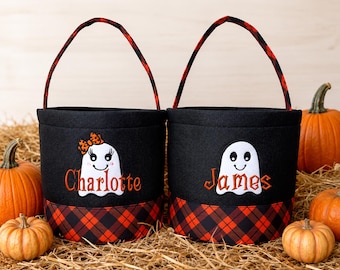 May include: Two black fabric buckets with red and black plaid trim and handles. Each bucket features an embroidered white ghost and a personalized name in orange thread. One ghost wears an orange leopard print bow. The names are Charlotte and James.