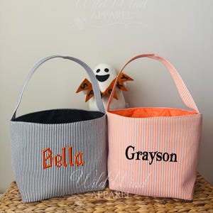 May include: Two striped fabric tote bags with handles. One is navy and white with the name "Bella" embroidered in orange. The other is peach and white with the name "Grayson" embroidered in black. A small ghost decoration is in the background.