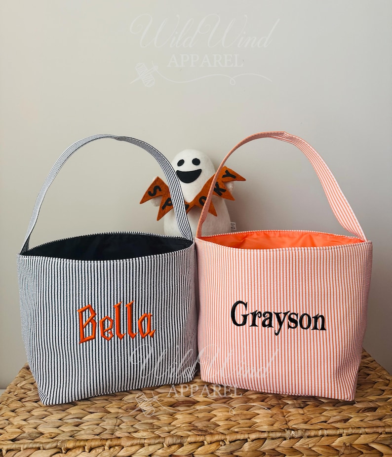 May include: Two striped fabric tote bags with handles. One is navy and white with the name "Bella" embroidered in orange. The other is peach and white with the name "Grayson" embroidered in black. A small ghost decoration is in the background.