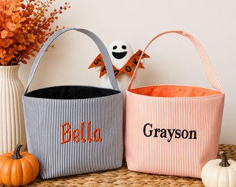 May include: Two fabric buckets with handles, one navy blue and white striped with the name Bella embroidered in orange, and one orange and white striped with the name Grayson embroidered in black. Both have open tops.