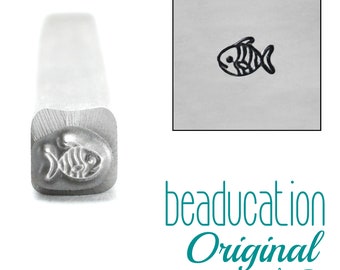 May include: A silver metal stamping tool with a square shaft and a fish design on the end. The tool creates a stamped impression of a small fish with striped markings and a circular eye. The text beaducation Original is visible in teal.