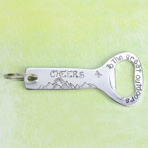 May include: A silver-toned metal bottle opener keychain featuring the engraved text "CHEERS" and "to the great outdoors." The design includes a mountain range with a rising sun, two small butterflies, and a circular metal ring attached to one end.