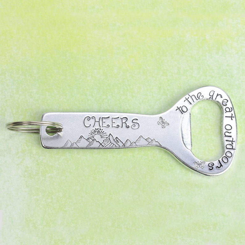 May include: A silver-toned metal bottle opener keychain featuring the engraved text "CHEERS" and "to the great outdoors." The design includes a mountain range with a rising sun, two small butterflies, and a circular metal ring attached to one end.