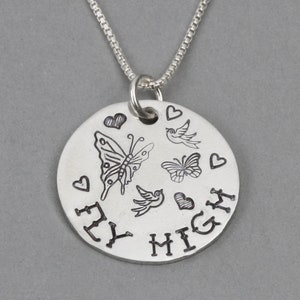May include: A silver-toned circular pendant necklace featuring an engraved design of two butterflies, two birds, and four hearts. The text FLY HIGH is stamped in black along the bottom edge of the disc, which hangs from a thin silver-toned box chain.