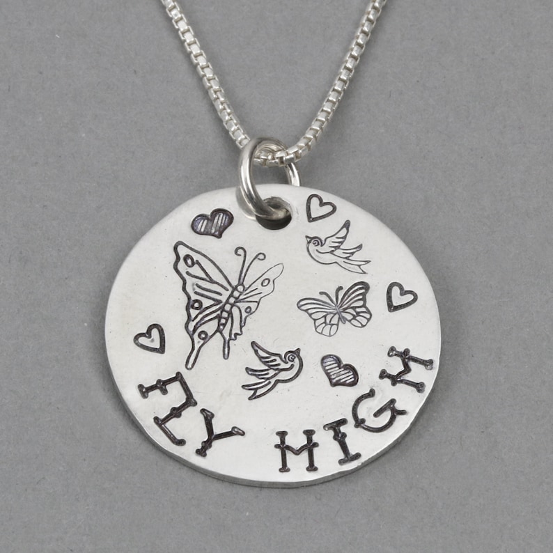 May include: A silver-toned circular pendant necklace featuring an engraved design of two butterflies, two birds, and four hearts. The text FLY HIGH is stamped in black along the bottom edge of the disc, which hangs from a thin silver-toned box chain.