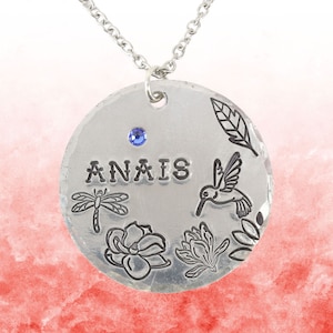 May include: A round silver pendant necklace featuring the stamped name ANAIS, a small blue crystal, and engraved illustrations of a dragonfly, a hummingbird, two flowers, and a leaf. The pendant hangs from a simple silver chain link necklace.
