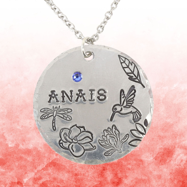 May include: A round silver pendant necklace featuring the stamped name ANAIS, a small blue crystal, and engraved illustrations of a dragonfly, a hummingbird, two flowers, and a leaf. The pendant hangs from a simple silver chain link necklace.