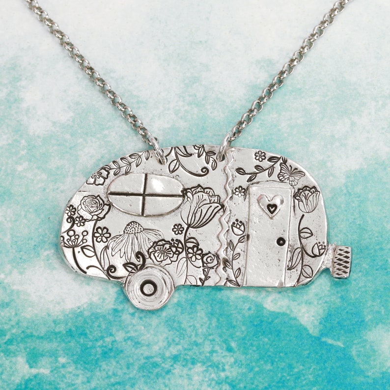 May include: A silver-toned pendant shaped like a vintage camper trailer, suspended from a matching metal chain. The surface features engraved floral patterns, a window with a cross design, and a door with a small heart cutout detail.