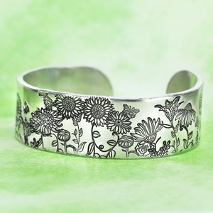 May include: A silver-toned metal cuff bracelet featuring a detailed black engraved pattern of various wildflowers, including sunflowers and daisies, interspersed with small butterflies across the entire outer surface of the band.