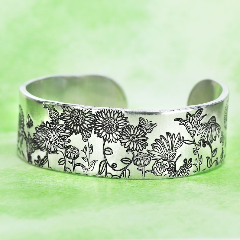 May include: A silver-toned metal cuff bracelet featuring a detailed black engraved pattern of various wildflowers, including sunflowers and daisies, interspersed with small butterflies across the entire outer surface of the band.