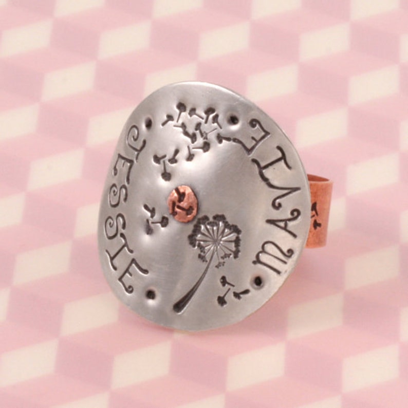 May include: A silver-toned metal ring featuring a circular face stamped with the names Jessie and Mavie, a dandelion flower, and floating seeds. A small copper-colored circular accent is centered on the face, and the ring band is copper.