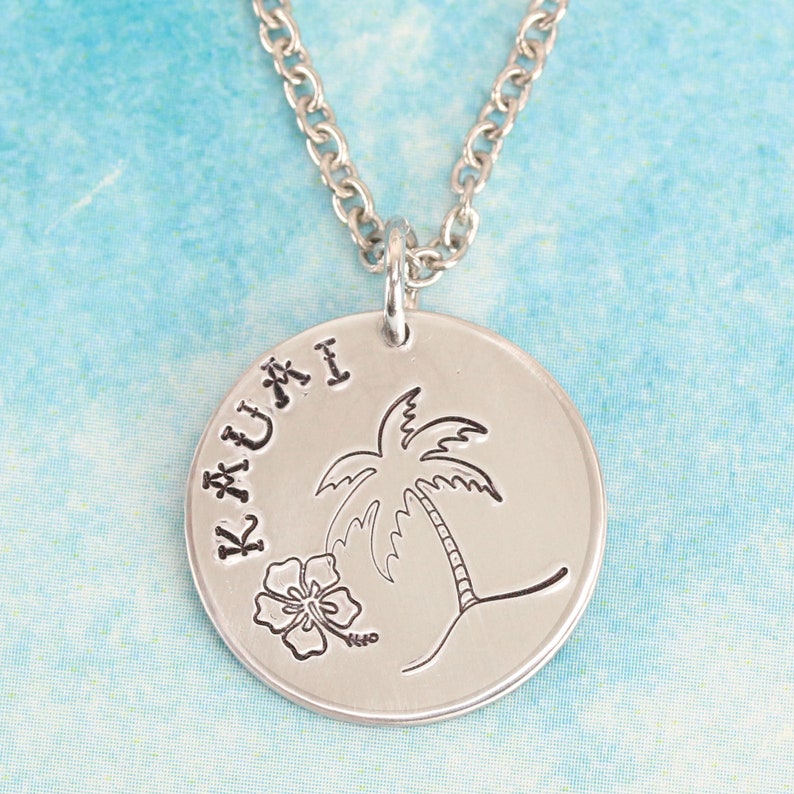 May include: A silver-toned circular pendant necklace featuring an engraved palm tree, a hibiscus flower, and the text KAUAI stamped along the left edge. The pendant hangs from a matching silver-toned metal chain with circular links.