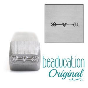 May include: A silver metal stamping tool with a rectangular shaft and a brushed finish. The bottom end features a recessed design of an arrow with a heart at its center. A square inset shows the stamped impression of the arrow and heart design.