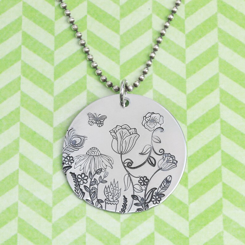 May include: A circular silver pendant necklace featuring a detailed black line art engraving of various flowers, a small butterfly, and a peacock feather. The pendant hangs from a silver-toned ball chain, showcasing a botanical garden design.
