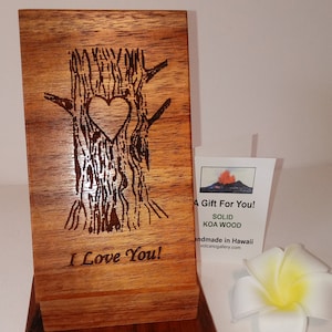 May include: A wooden sign with a heart carved into a tree trunk. The sign reads "I Love You!" and is made of solid Koa wood. A small card with the text "A Gift For You! Solid Koa Wood Handmade in Hawaii volcanogallery.com" is next to the sign.