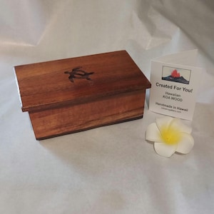 May include: A rectangular wooden box with a turtle design on the lid. The box is made of Koa wood and has a rich brown color. A white flower and a card that says "Created For You! Hawaiian KOA WOOD" are next to the box.