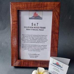 May include: A rectangular picture frame crafted from reddish-brown solid Koa wood with a visible grain. The frame holds a white insert with the text 5 x 7 SOLID KOA WOOD FRAME Made in Volcano, Hawaii. A white plumeria flower sits in front.