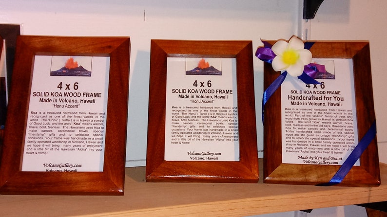 May include: Three 4x6 inch solid Koa wood picture frames with a blue ribbon and a white flower on the rightmost frame. The frames are made in Volcano, Hawaii and feature a volcano image and text about Koa wood.