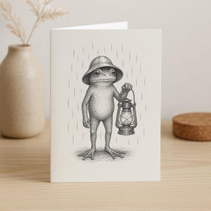 May include: A greeting card with a detailed illustration of a frog wearing a hat and holding a lantern in the rain. The frog is depicted in a realistic style with intricate shading, and the lantern has a classic design. The background shows vertical lines representing rainfall.