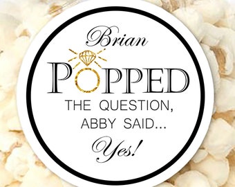 Pode incluir: Um saco de plástico transparente cheio de pipocas brancas, atado no topo com uma fita de cetim preta. Uma etiqueta redonda branca apresenta o texto Brian POPPED THE QUESTION, ABBY SAID... Yes! com um gráfico de anel de diamante dourado.
