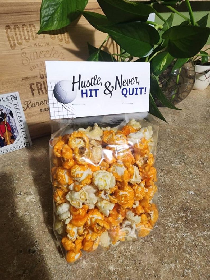 Pode incluir: Um saco de pl&aacute;stico transparente cheio de pipocas cor de laranja e brancas, selado com um topo de papel branco com um gr&aacute;fico de uma bola de voleibol e o texto preto "Hustle, HIT & Never, QUIT!" em fontes script e sem serifa.