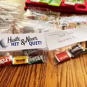 Pode incluir: Sacos de pl&aacute;stico transparentes cheios de barras de chocolate em miniatura sortidas, cada um com um cabe&ccedil;alho de papel branco. O cabe&ccedil;alho esquerdo apresenta um gr&aacute;fico de voleibol e o texto Hustle, & Never, HIT & QUIT! O direito diz Good luck Grammys K.