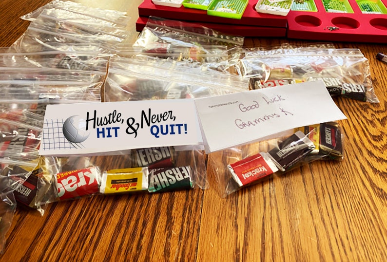 Pode incluir: Sacos de pl&aacute;stico transparentes cheios de barras de chocolate em miniatura sortidas, cada um com um cabe&ccedil;alho de papel branco. O cabe&ccedil;alho esquerdo apresenta um gr&aacute;fico de voleibol e o texto Hustle, & Never, HIT & QUIT! O direito diz Good luck Grammys K.