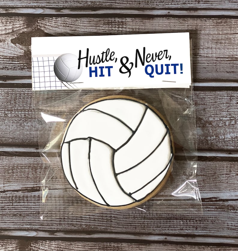 Pode incluir: Um biscoito redondo decorado com glac&ecirc; branco e linhas pretas para se parecer com uma bola de v&ocirc;lei, embalado em um saco pl&aacute;stico transparente com um topo de papel branco com um gr&aacute;fico de v&ocirc;lei e o texto Hustle, & Never, HIT & QUIT!