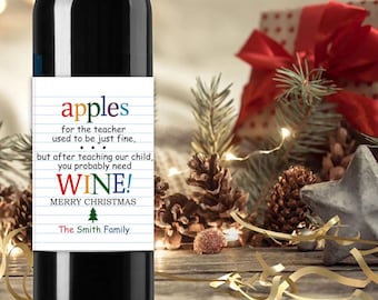 Puede incluir: Una botella de vino de vidrio oscuro con una etiqueta rectangular blanca que presenta gráficos de papel con líneas azules. El texto dice: apples for the teacher used to be just fine, but after teaching our child, you probably need WINE! MERRY CHRISTMAS.