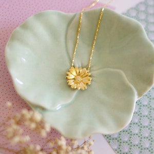 May include: A gold necklace with a delicate chain and a flower pendant. The pendant is a detailed design of a daisy with a yellow gold finish. The necklace is displayed on a light green ceramic dish.