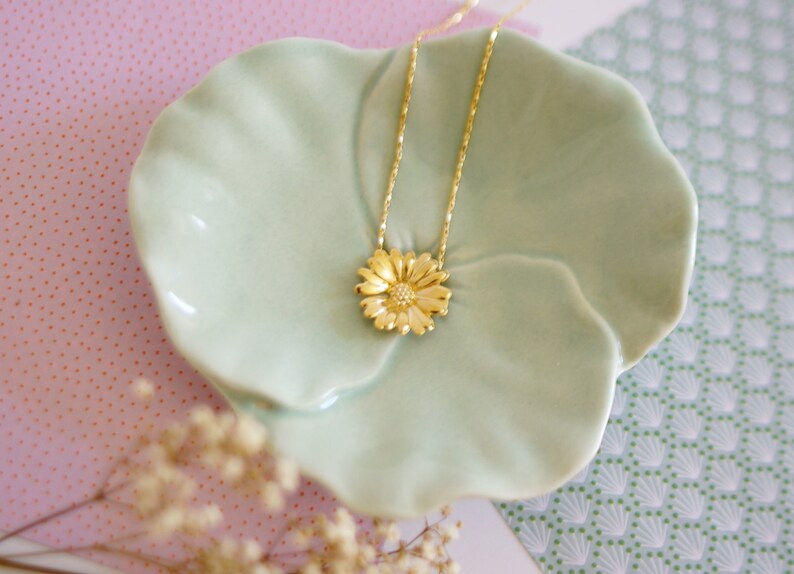 May include: A gold necklace with a delicate chain and a flower pendant. The pendant is a detailed design of a daisy with a yellow gold finish. The necklace is displayed on a light green ceramic dish.