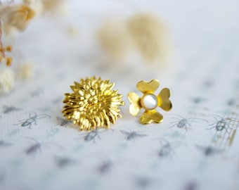 May include: Two gold-colored earrings. One is a detailed flower shape, and the other is a four-leaf clover design with a pearl center. The earrings are on a patterned surface with a soft focus background.
