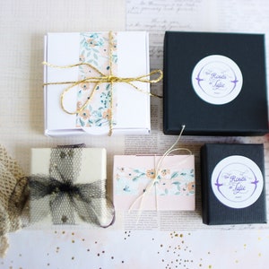 May include: Four small gift boxes with floral patterned paper and black and white ribbon. The boxes are on a white surface with a white and gold patterned background. The boxes have a logo that says "The Roses of Julie" in purple text.