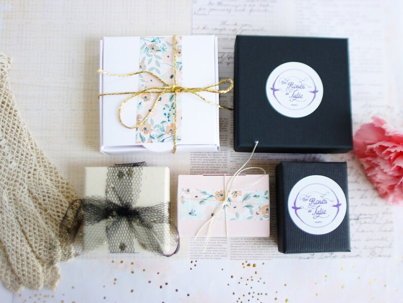 May include: Four small gift boxes with floral patterned paper and black and white ribbon. The boxes are on a white surface with a white and gold patterned background. The boxes have a logo that says "The Roses of Julie" in purple text.
