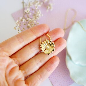 May include: A gold-toned necklace with a delicate chain and a flower pendant. The flower pendant is a detailed design with a textured surface.