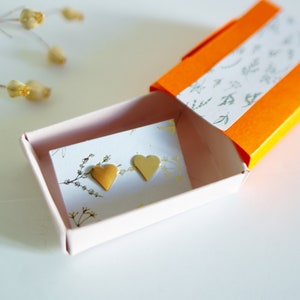 May include: A pair of gold heart-shaped earrings in a small pink box with a white card inside. The box is open and the earrings are visible. The box is sitting on a white surface.