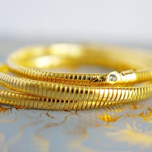 Op de afbeelding: Een opgerolde goudkleurige armband met een slangachtig ontwerp. De armband is gemaakt van gesegmenteerde metalen schakels en heeft een kleine, heldere edelsteen bij de sluiting. De sieraden worden gepresenteerd op een patroonoppervlak.