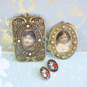 May include: Two antique brass picture frames, one rectangular and one oval, each holding a vintage photograph. Below, a pair of oval earrings with floral designs. The frames and earrings are on a light blue surface.