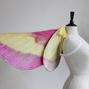 Pu&ograve; includere: Un paio di ali di falena caratterizzate da una sfumatura di colori rosa vivace e giallo tenue. Le ali sono fissate a un'imbracatura elastica bianca e presentano un bordo giallo testurizzato e soffice alla base, vicino alle spalline.