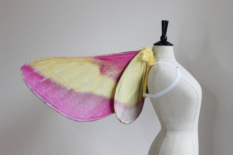Pu&ograve; includere: Un paio di ali di falena caratterizzate da una sfumatura di colori rosa vivace e giallo tenue. Le ali sono fissate a un'imbracatura elastica bianca e presentano un bordo giallo testurizzato e soffice alla base, vicino alle spalline.